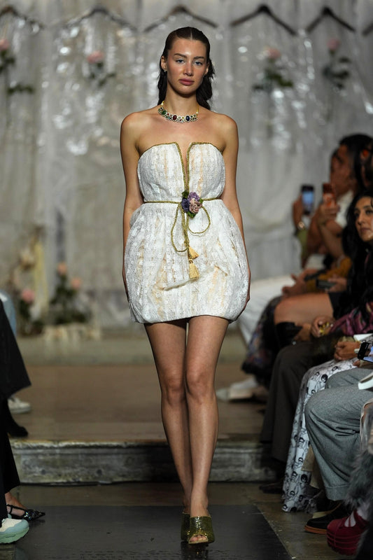 A model walks the runway wearing a strapless white textured mini dress with gold trim and a tassel tie at the waist, photographed during a fashion show with seated spectators in the background.