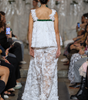 A model viewed from behind walking down a runway wearing a white lace sleeveless dress with ruffled straps, a green band at the back, and a long sheer lace skirt.