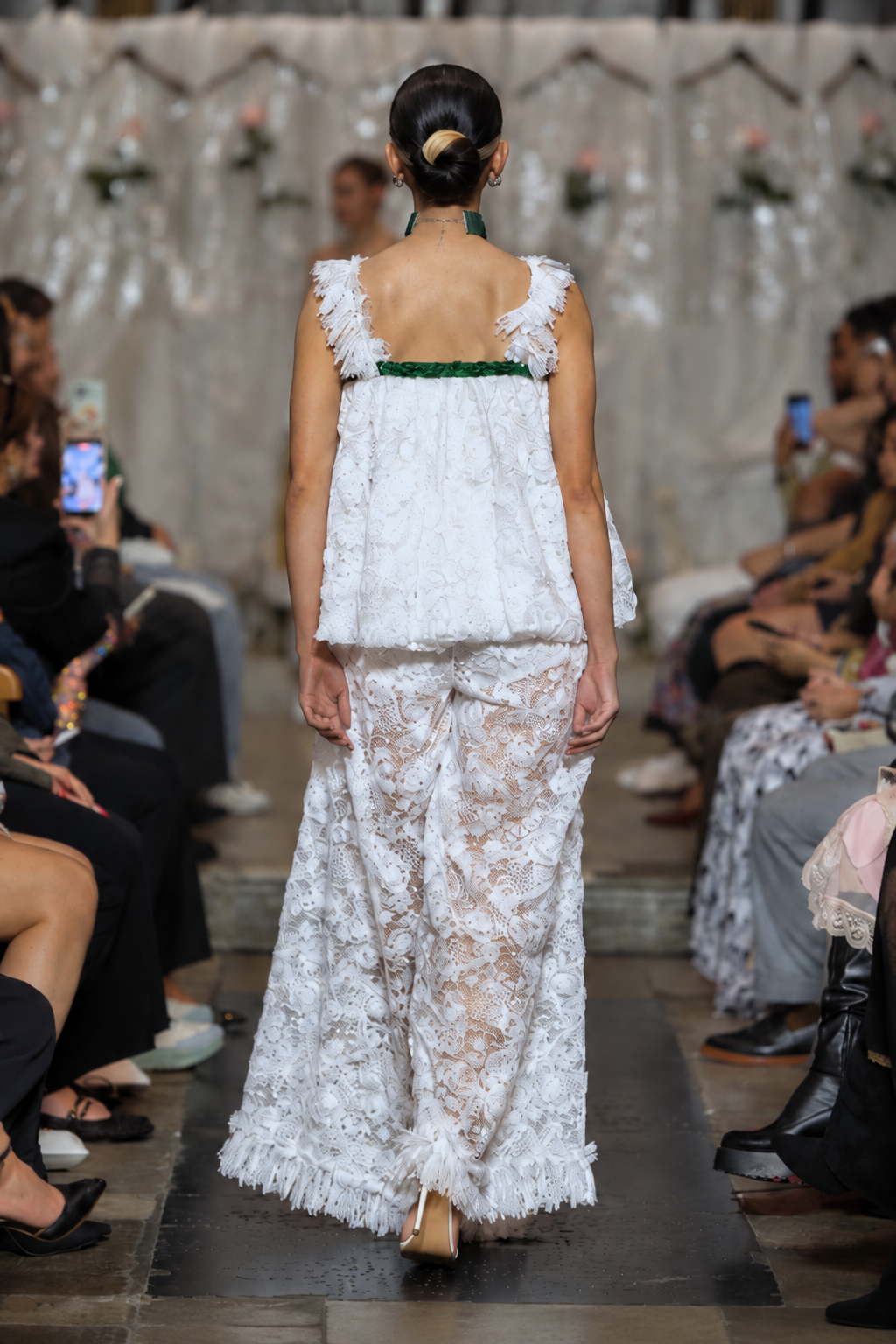 A model viewed from behind walking down a runway wearing a white lace sleeveless dress with ruffled straps, a green band at the back, and a long sheer lace skirt.