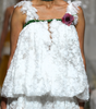 A model walks a fashion runway wearing a white textured lace two-piece outfit with a ruffled top and wide-leg pants, accessorized with a dark green collar and a floral chest detail. Audience members are seated on both sides taking photos.