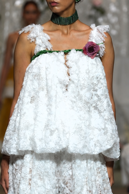 A model walks a fashion runway wearing a white textured lace two-piece outfit with a ruffled top and wide-leg pants, accessorized with a dark green collar and a floral chest detail. Audience members are seated on both sides taking photos.