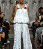 A model walks a fashion runway wearing a white textured lace two-piece outfit with a ruffled top and wide-leg pants, accessorized with a dark green collar and a floral chest detail. Audience members are seated on both sides taking photos.