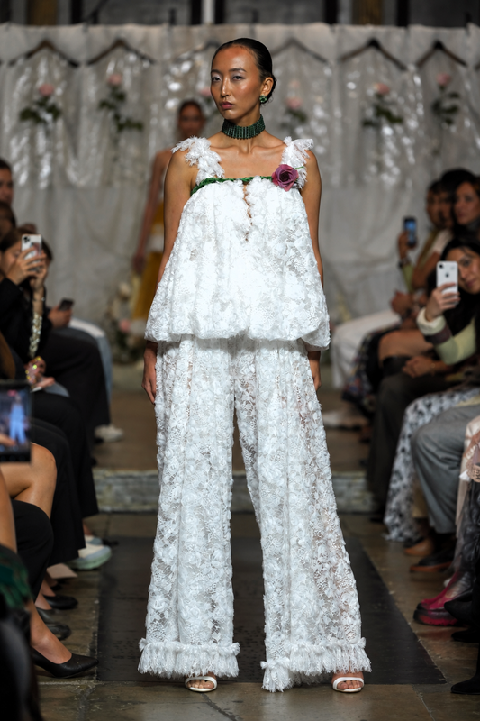 A model walks a fashion runway wearing a white textured lace two-piece outfit with a ruffled top and wide-leg pants, accessorized with a dark green collar and a floral chest detail. Audience members are seated on both sides taking photos.