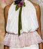 A runway photo of a model walking down a fashion show catwalk wearing a white halter top with a pink ruffled overlay, sheer white tights with lace trim, and a green and purple floral neck accent.