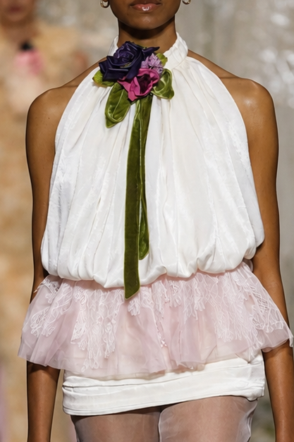 A runway photo of a model walking down a fashion show catwalk wearing a white halter top with a pink ruffled overlay, sheer white tights with lace trim, and a green and purple floral neck accent.