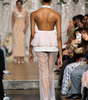 A model viewed from behind walking down a fashion runway wearing a sheer light pink and white backless dress with a ruffled waist and flared tulle legs. Audience members sit along the sides of the runway.