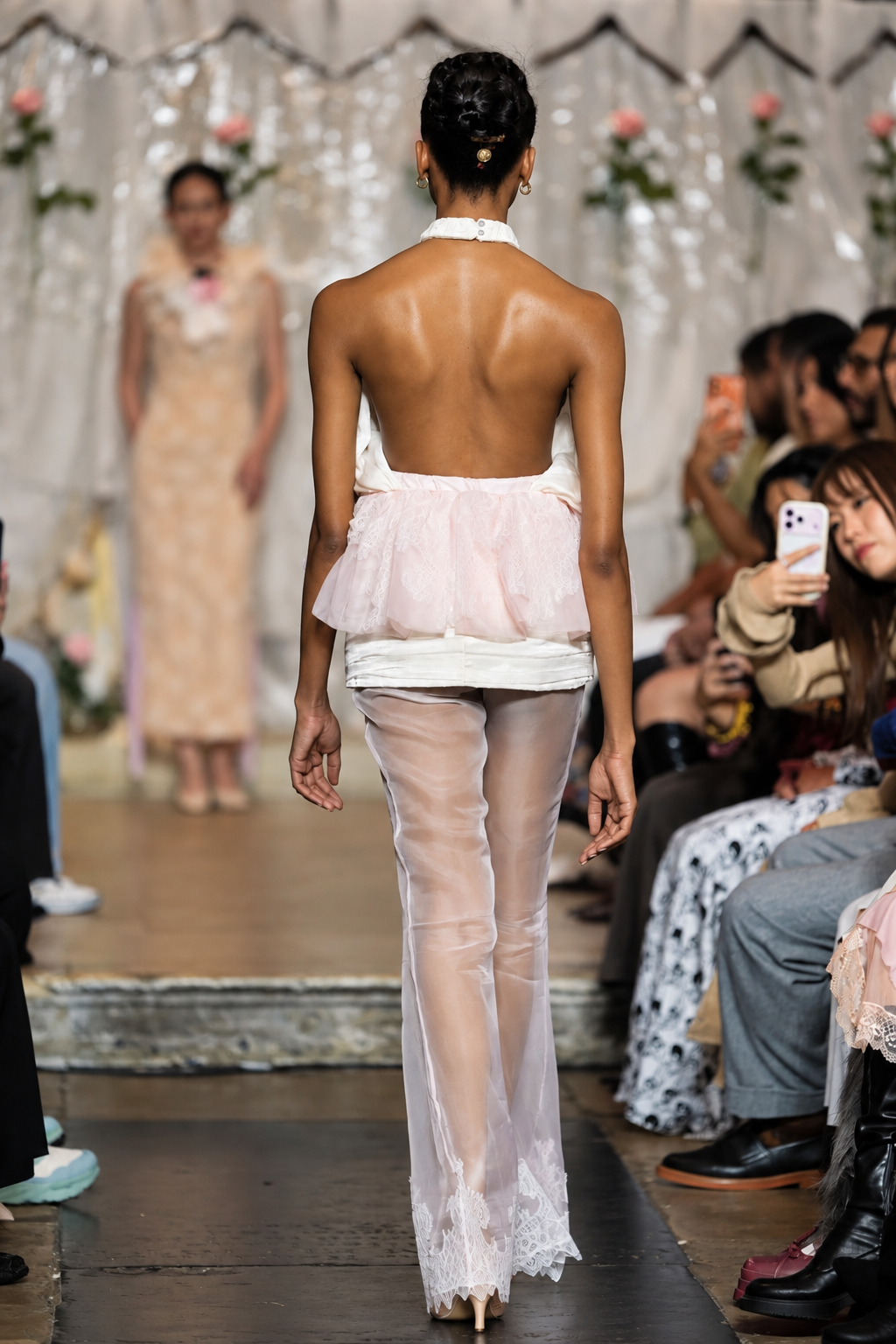 A model viewed from behind walking down a fashion runway wearing a sheer light pink and white backless dress with a ruffled waist and flared tulle legs. Audience members sit along the sides of the runway.