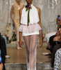 A runway photo of a model walking down a fashion show catwalk wearing a white halter top with a pink ruffled overlay, sheer white tights with lace trim, and a green and purple floral neck accent.