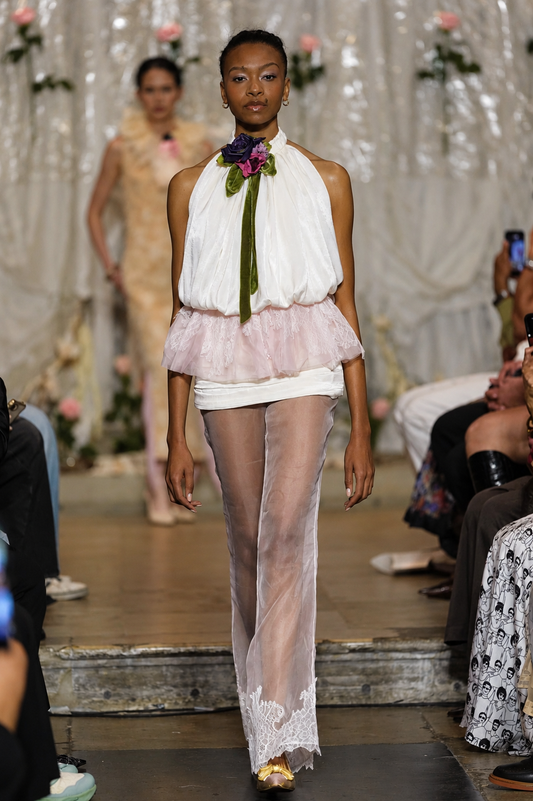 A runway photo of a model walking down a fashion show catwalk wearing a white halter top with a pink ruffled overlay, sheer white tights with lace trim, and a green and purple floral neck accent.