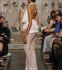 A model walks down a fashion runway wearing a sheer, light pink and white halter dress with a ruffled tulle overlay and lace-trimmed hem. Audience members are seated on both sides of the runway.