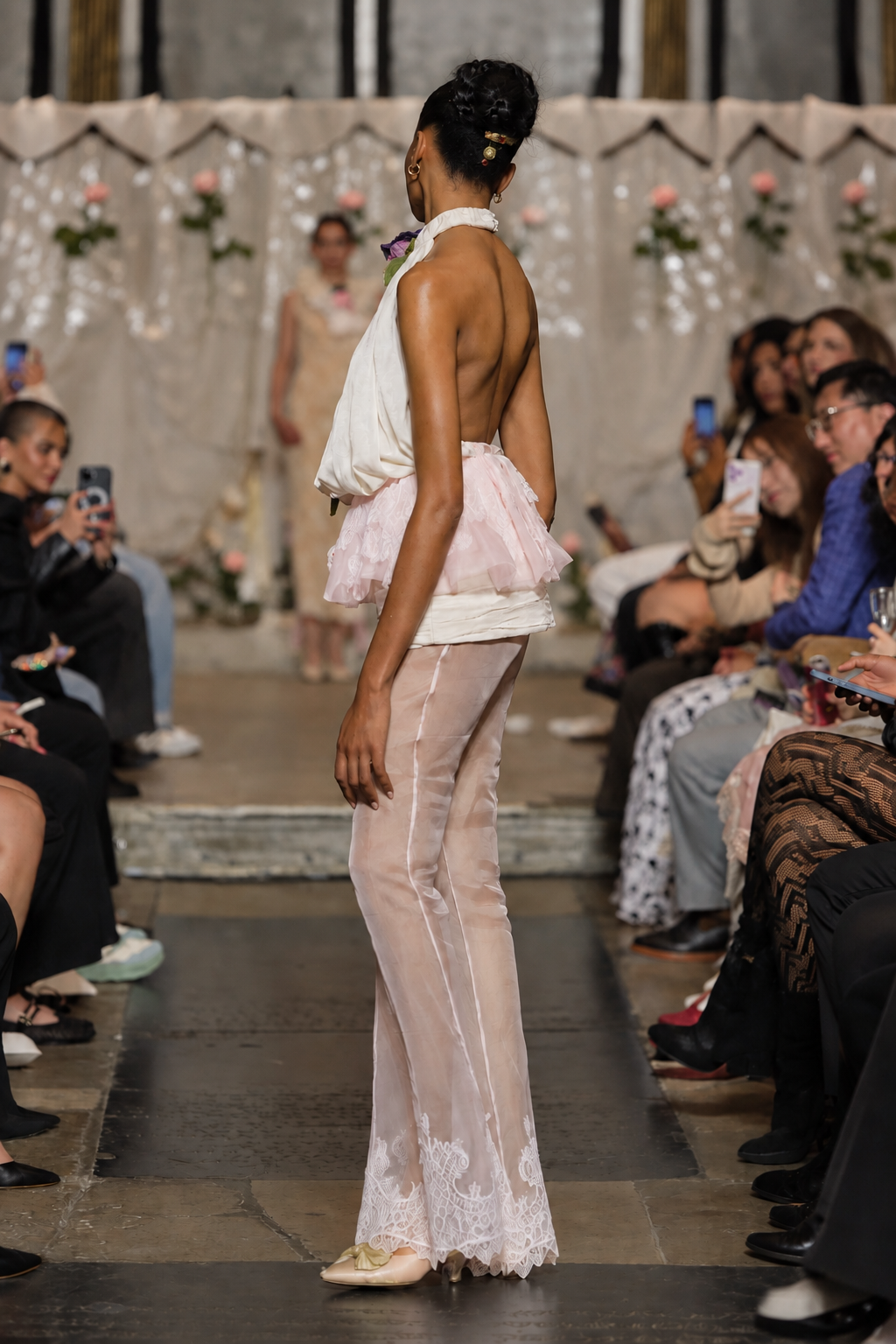 A model walks down a fashion runway wearing a sheer, light pink and white halter dress with a ruffled tulle overlay and lace-trimmed hem. Audience members are seated on both sides of the runway.