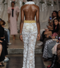 A model shown from behind on a fashion runway wearing a white floral lace halter jumpsuit with a gold waistband and feather trim at the neckline and hem. The runway is surrounded by an audience.