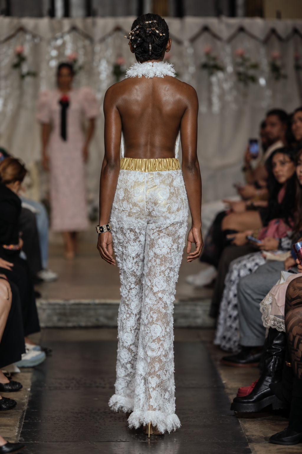 A model shown from behind on a fashion runway wearing a white floral lace halter jumpsuit with a gold waistband and feather trim at the neckline and hem. The runway is surrounded by an audience.