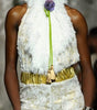 A model walks the runway wearing a white textured lace and faux-fur outfit with a gold belt and a bright green tie detail. The photo is taken on a fashion show with an audience seated in the background.