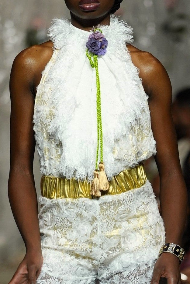 A model walks the runway wearing a white textured lace and faux-fur outfit with a gold belt and a bright green tie detail. The photo is taken on a fashion show with an audience seated in the background.