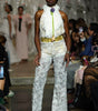 A model walks the runway wearing a white textured lace and faux-fur outfit with a gold belt and a bright green tie detail. The photo is taken on a fashion show with an audience seated in the background.