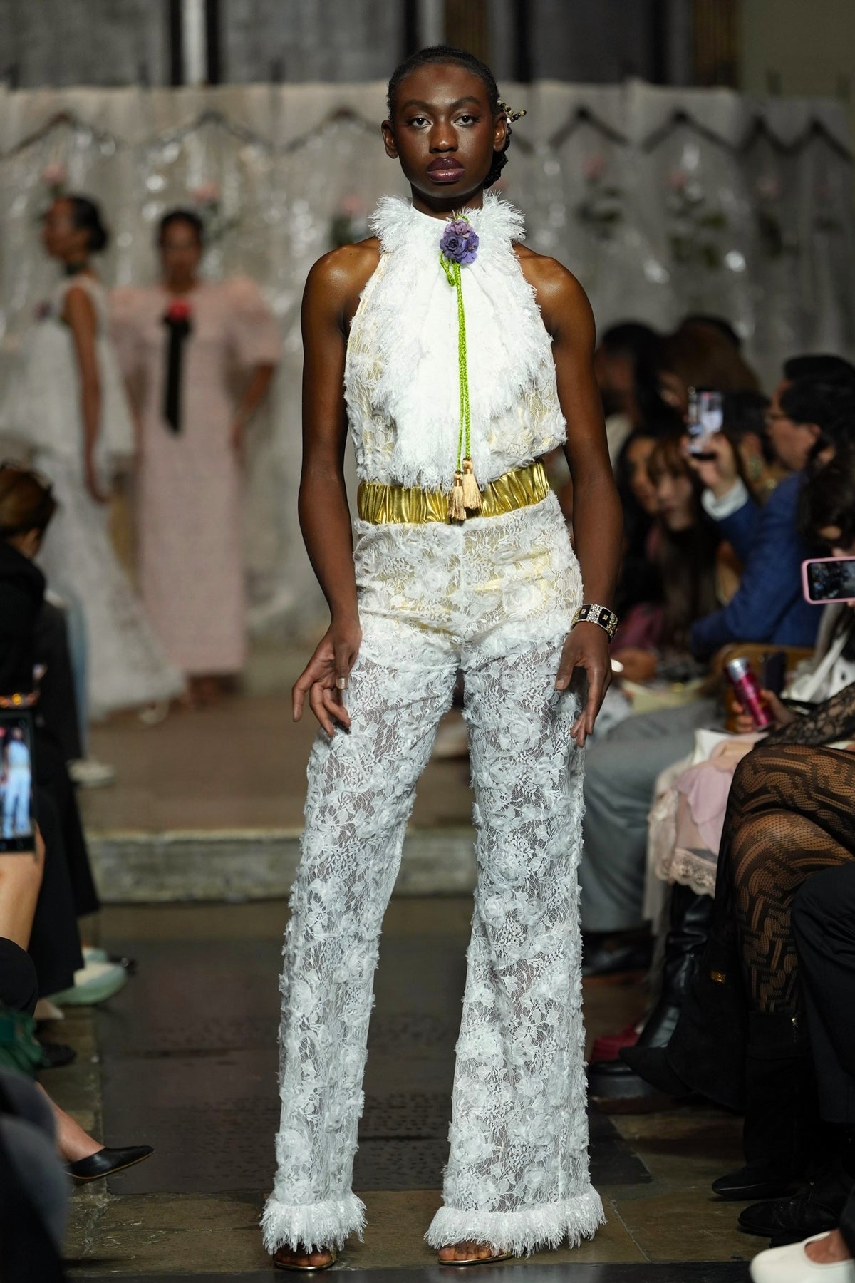 A model walks the runway wearing a white textured lace and faux-fur outfit with a gold belt and a bright green tie detail. The photo is taken on a fashion show with an audience seated in the background.