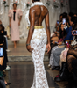 A model on a fashion runway viewed from behind, wearing a white lace floor-length gown with feather trim and a gold waistband, with a seated audience watching on the side.
