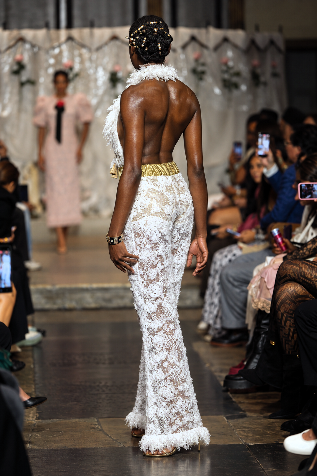 A model on a fashion runway viewed from behind, wearing a white lace floor-length gown with feather trim and a gold waistband, with a seated audience watching on the side.