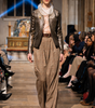 Model walking on a fashion show runway wearing a metallic jacket and plaid pants.