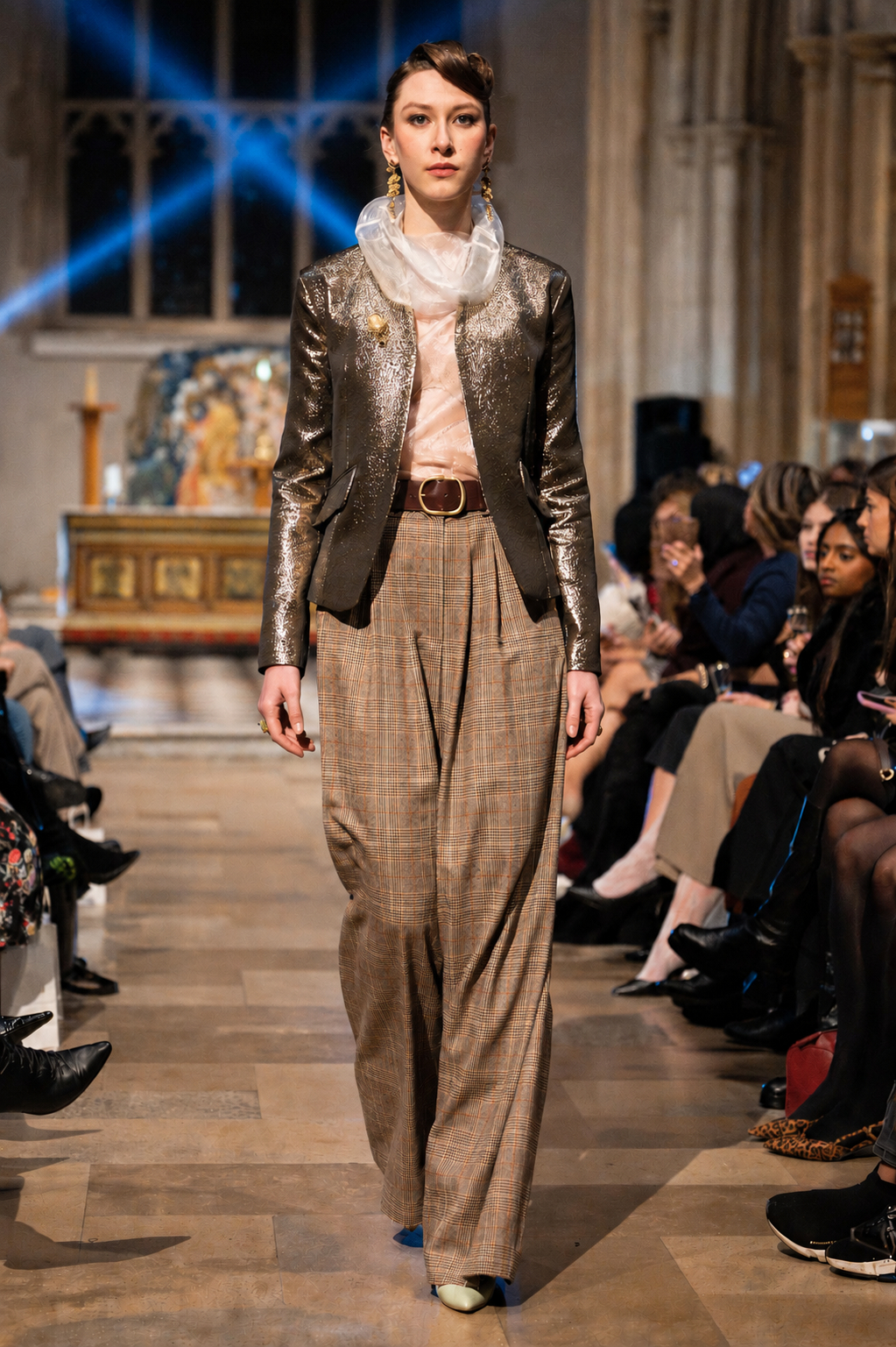 Model walking on a fashion show runway wearing a metallic jacket and plaid pants.
