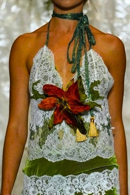 A model walks a fashion runway wearing a halter-neck mini dress made of white lace and green fabric, decorated with a red and yellow floral applique at the bust and a white fringed hem.