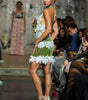 A model walks a fashion runway wearing a green and white lace halter mini dress with a ruffled hem and beige high heels, with an audience seated in the background.