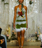 A model walks a fashion runway wearing a halter-neck mini dress made of white lace and green fabric, decorated with a red and yellow floral applique at the bust and a white fringed hem.