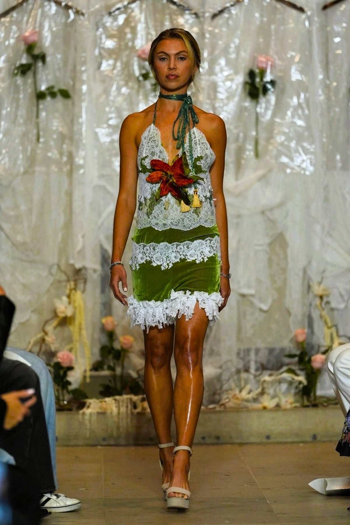 A model walks a fashion runway wearing a halter-neck mini dress made of white lace and green fabric, decorated with a red and yellow floral applique at the bust and a white fringed hem.