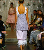 A model walks down a fashion runway wearing a lavender patterned sleeveless top with white ruffled shoulder straps and light pink ruffled pants, photographed from behind in a fashion show setting with seated spectators on both sides.