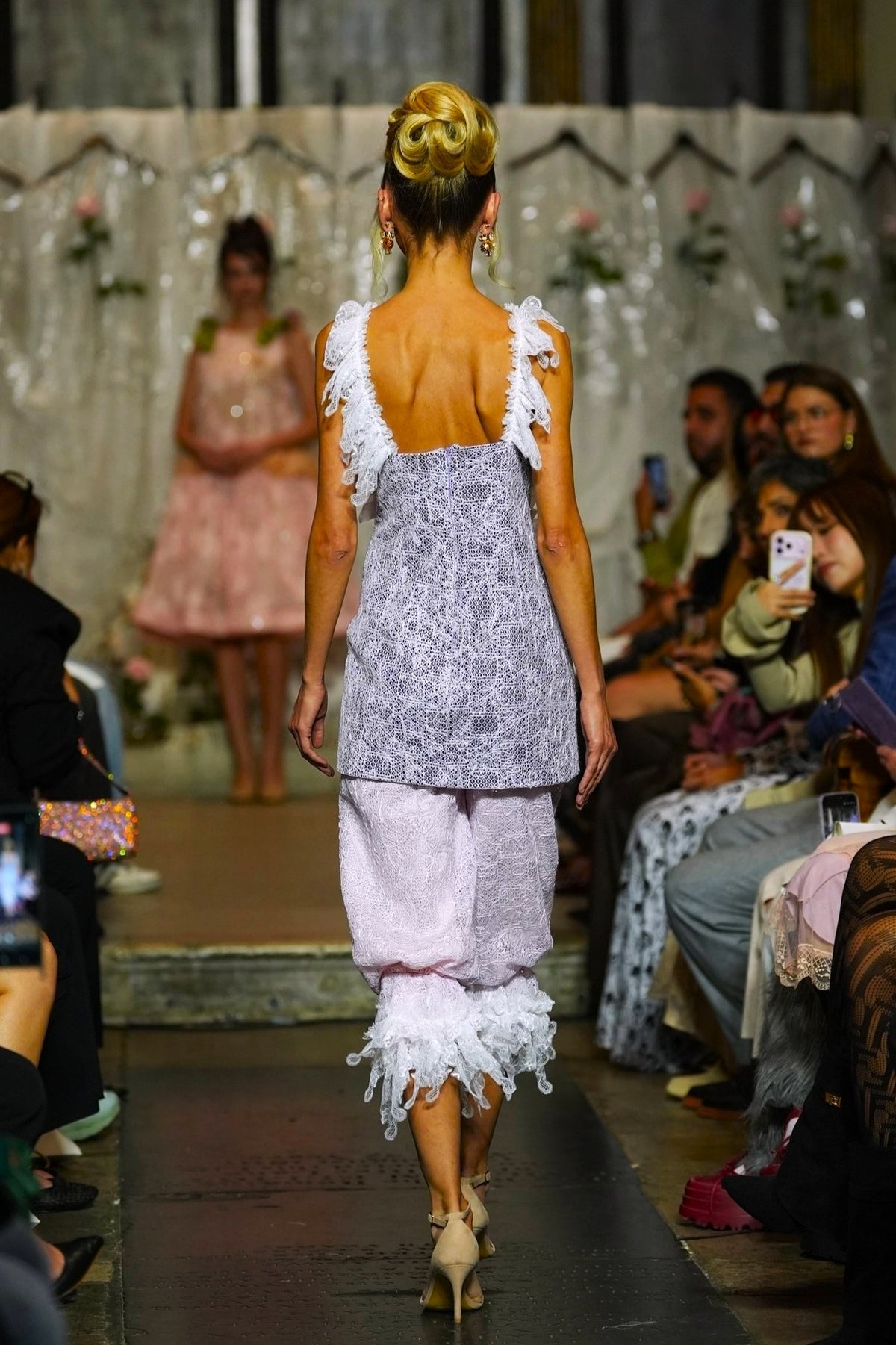 A model walks down a fashion runway wearing a lavender patterned sleeveless top with white ruffled shoulder straps and light pink ruffled pants, photographed from behind in a fashion show setting with seated spectators on both sides.