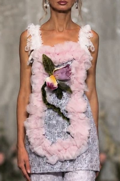 A model walks a fashion runway wearing a light pink and white embellished sleeveless top with a floral applique and matching ruffled cropped pants.