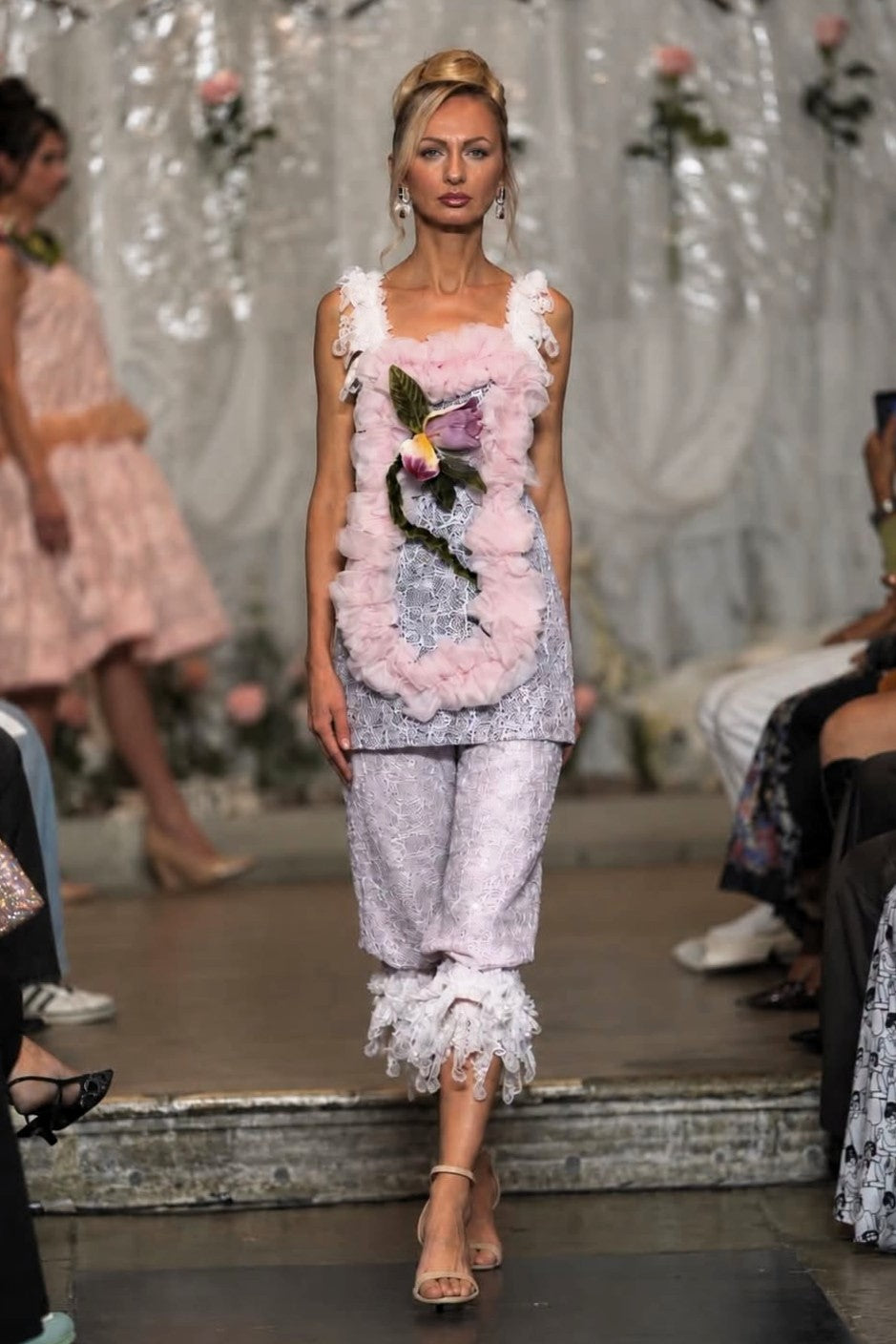 A model walks a fashion runway wearing a light pink and white embellished sleeveless top with a floral applique and matching ruffled cropped pants.