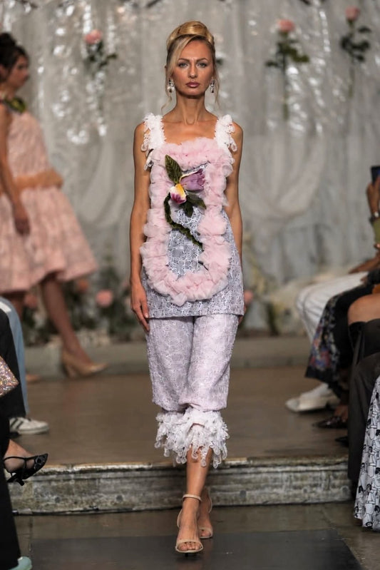 A model walks a fashion runway wearing a light pink and white embellished sleeveless top with a floral applique and matching ruffled cropped pants.