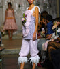 A model walks a fashion runway wearing a light lavender textured two-piece set with white ruffled trim, a fuzzy pink feathered vest, and beige high-heeled sandals. The background shows a fashion show setting with seated spectators and another model in the distance.