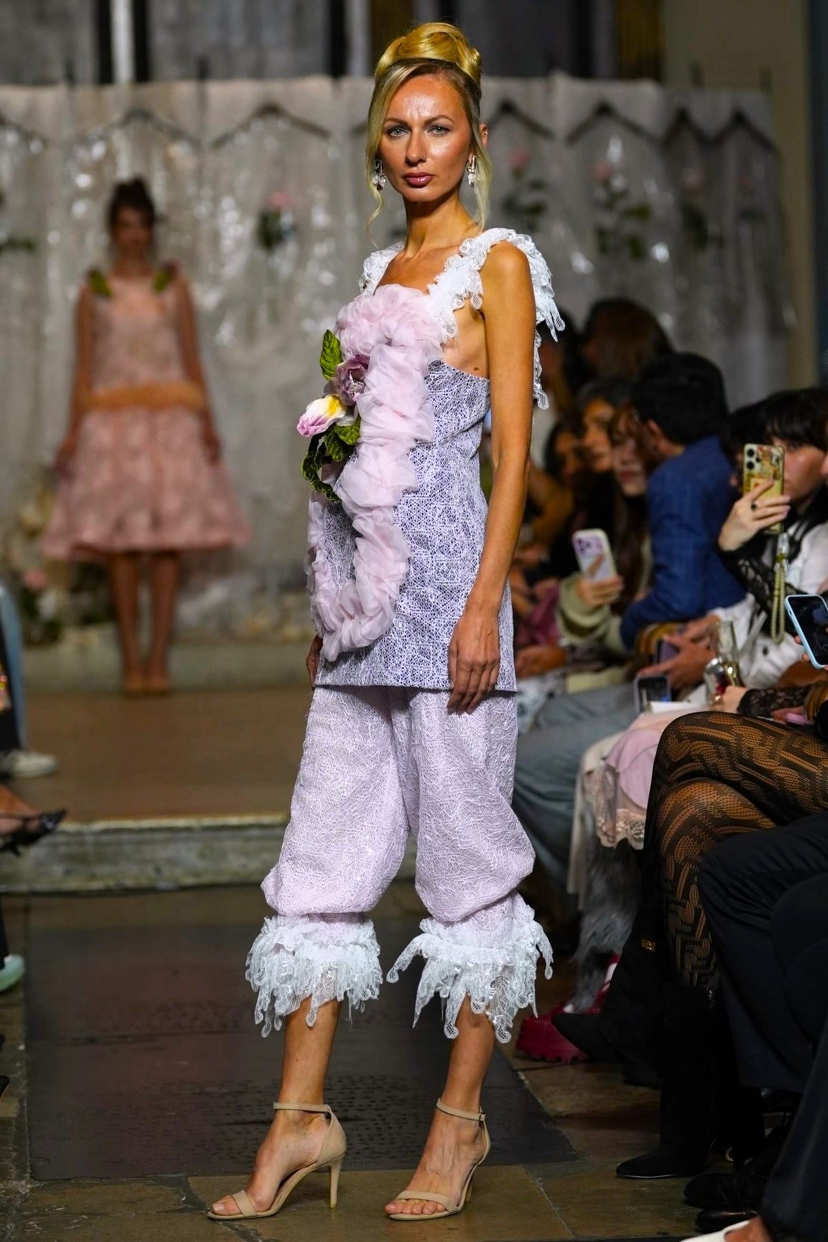 A model walks a fashion runway wearing a light lavender textured two-piece set with white ruffled trim, a fuzzy pink feathered vest, and beige high-heeled sandals. The background shows a fashion show setting with seated spectators and another model in the distance.