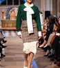 Model walking on a fashion show runway wearing a green top, plaid skirt, and white lace details.