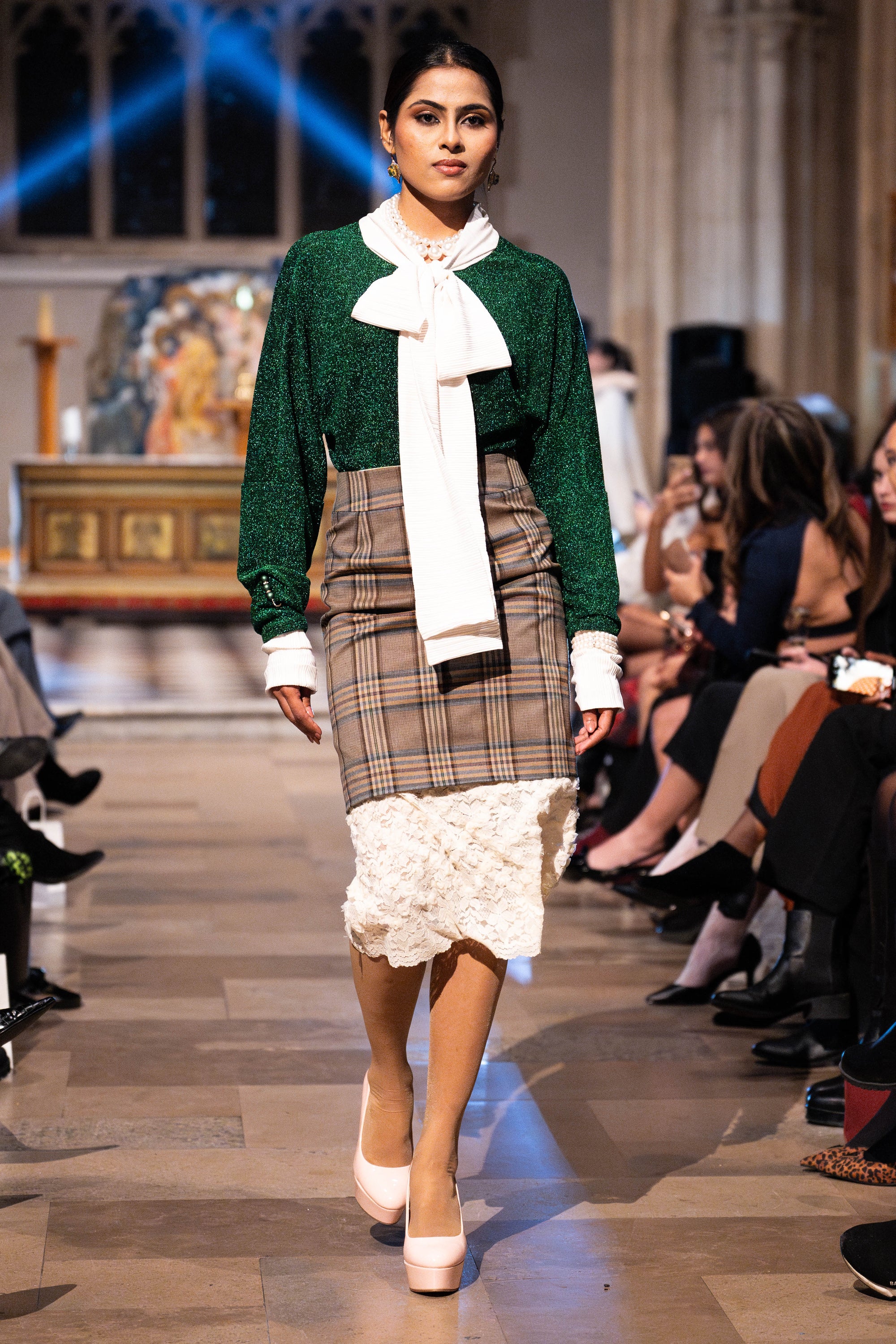 Model walking on a fashion show runway wearing a green top, plaid skirt, and white lace details.