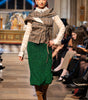 Model walking on a runway wearing a plaid top and green skirt with an audience watching.