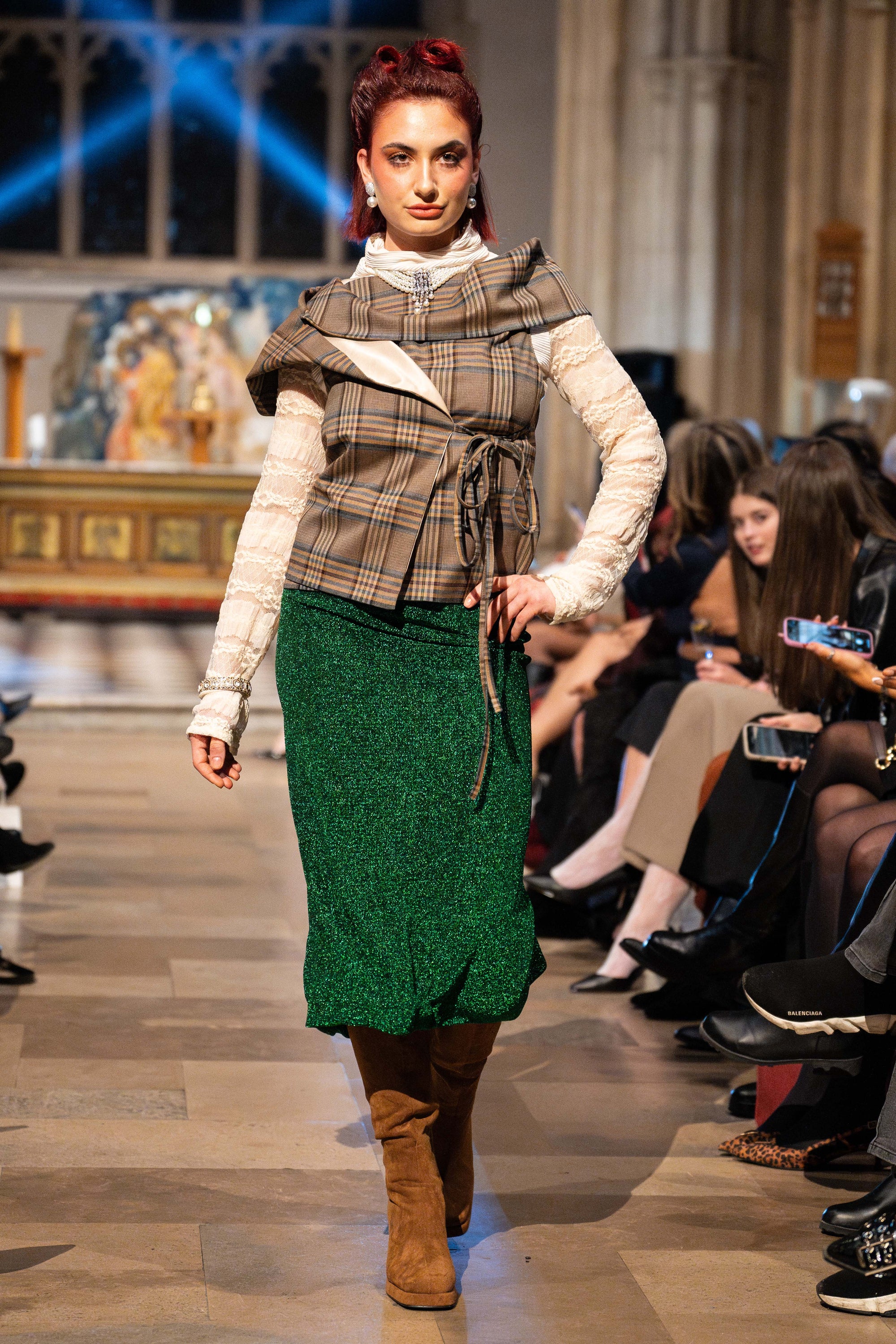Model walking on a runway wearing a plaid top and green skirt with an audience watching.
