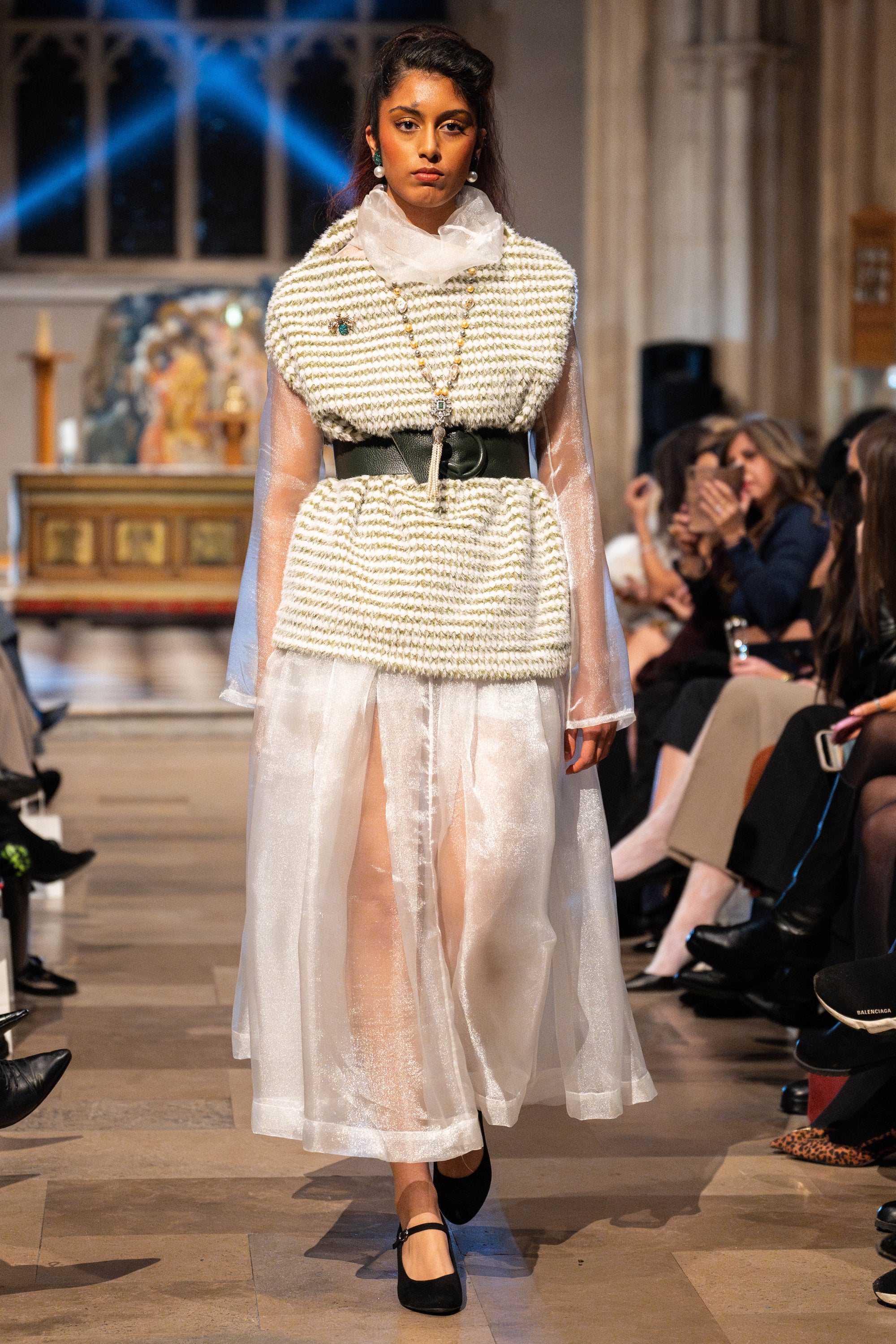 Model walking on a runway wearing a textured top and sheer skirt with a blurred audience in the background.