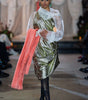 Model wearing a green and silver dress with a pink coat, standing on a runway.