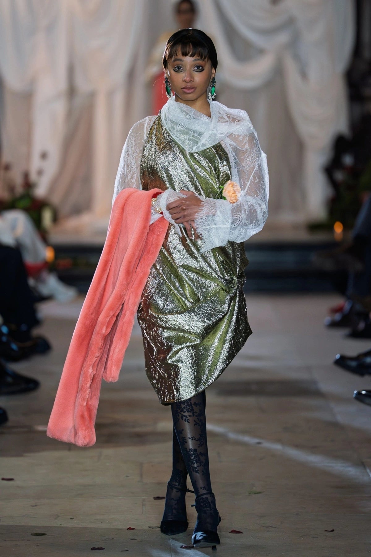Model wearing a green and silver dress with a pink coat, standing on a runway.