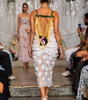 A model from behind wearing a white floral lace dress with green straps and a gold beaded back detail, walking down a fashion runway.
