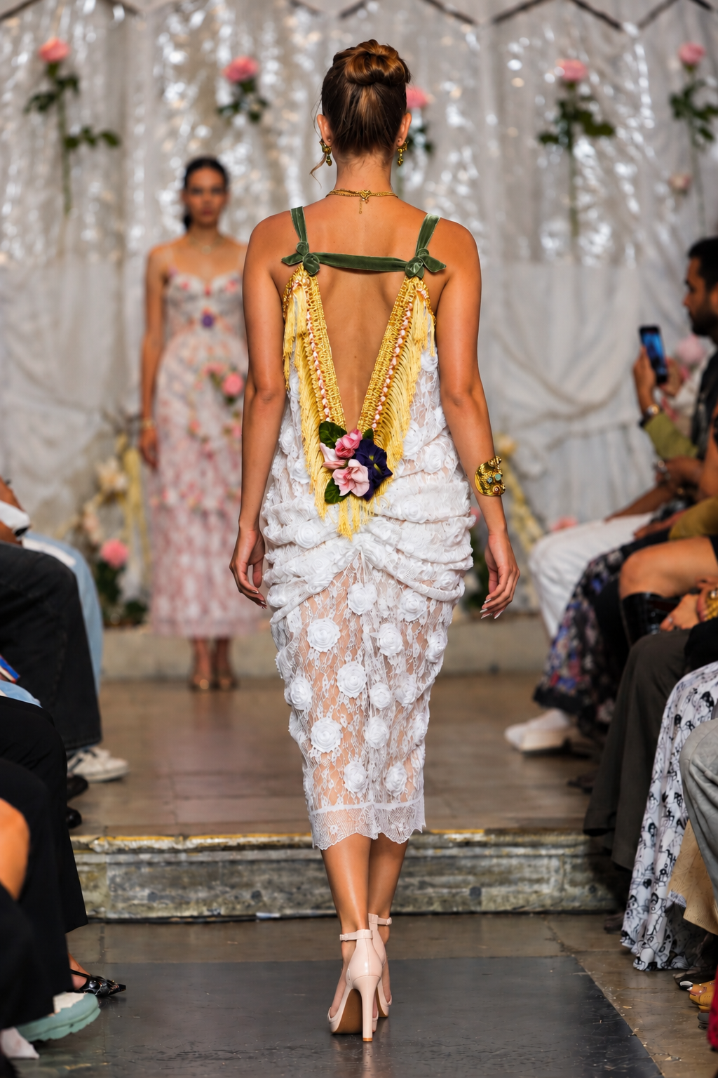 A model from behind wearing a white floral lace dress with green straps and a gold beaded back detail, walking down a fashion runway.