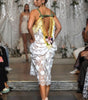 A model on a runway wearing a white floral lace dress with green and gold straps and a back flower detail, photographed from behind on a fashion show.