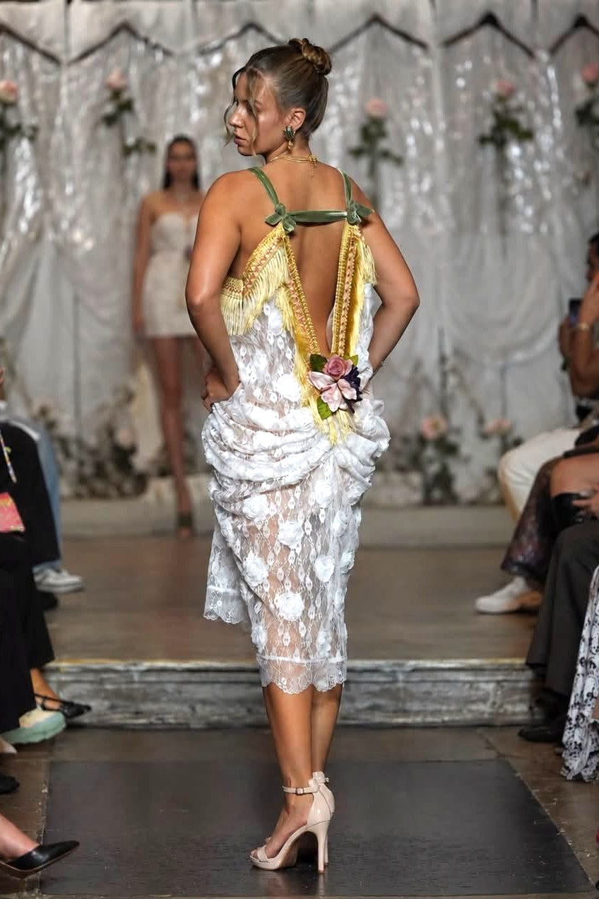 A model on a runway wearing a white floral lace dress with green and gold straps and a back flower detail, photographed from behind on a fashion show.