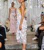 A model walks a fashion runway wearing a white floral lace midi dress with a yellow and green patterned halter top. The runway is decorated with white drapes and flowers, with audience members seated on both sides.