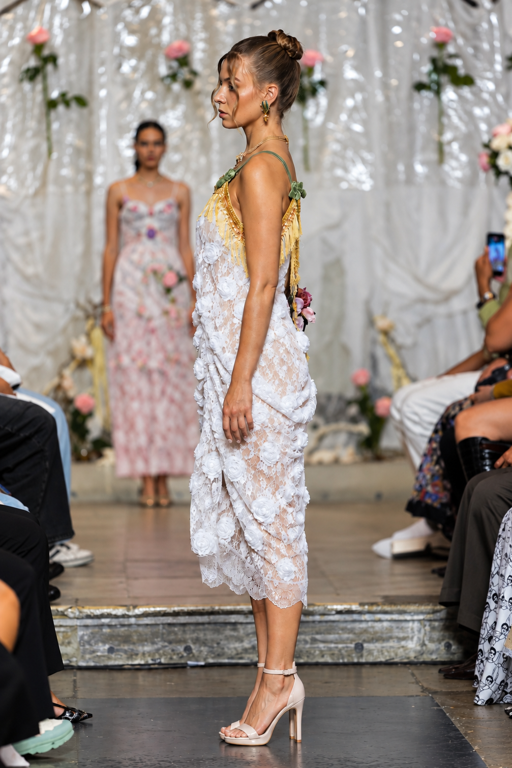 A model walks a fashion runway wearing a white floral lace midi dress with a yellow and green patterned halter top. The runway is decorated with white drapes and flowers, with audience members seated on both sides.