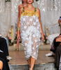 A model walks a fashion runway wearing a sheer white floral lace dress with a gold fringe top and green shoulder straps, set against a decorated white draped backdrop with flowers.